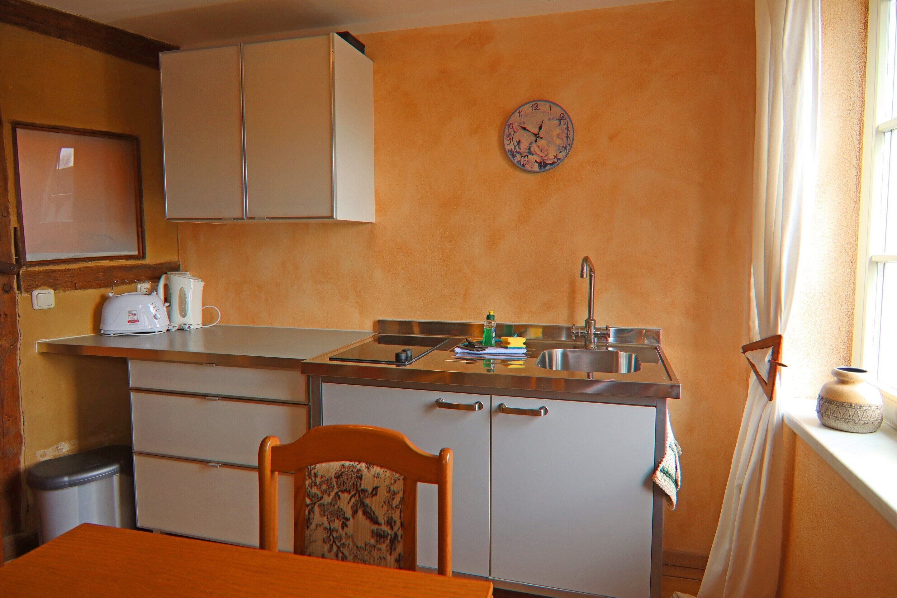 Una cocina compacta con un lavamanos, encimera y armarios. Se observa una silla y una mesa en primer plano.