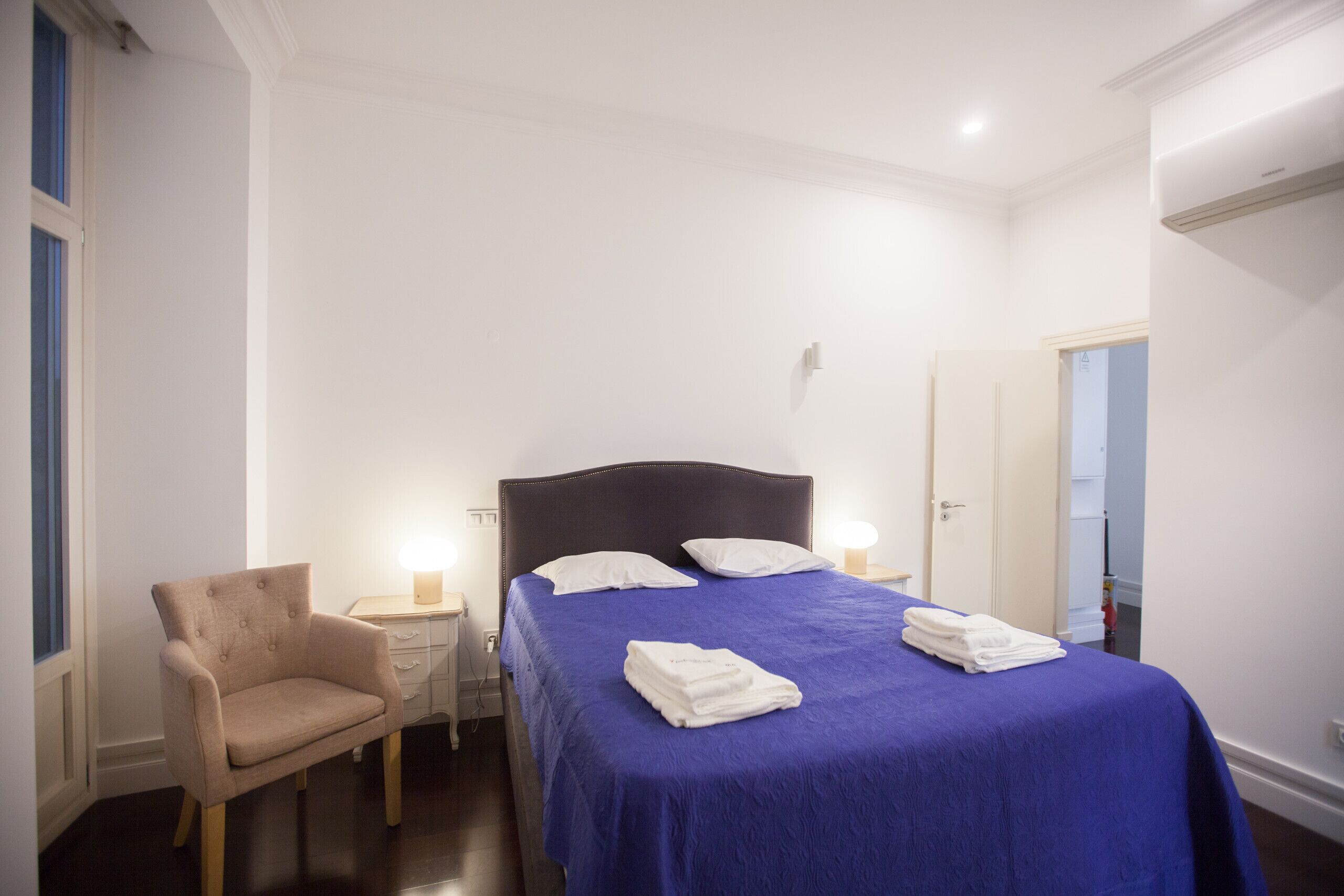 Una cama bien hecha con una colcha azul y almohadas blancas, una silla, una mesita de noche con una lámpara y una puerta que da a otra habitación.