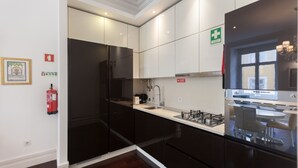 Una cocina moderna con armarios oscuros, un refrigerador de acero inoxidable, una estufa a gas y un lavamanos. Hay un extintor en la pared y un letrero de primeros auxilios sobre la puerta.