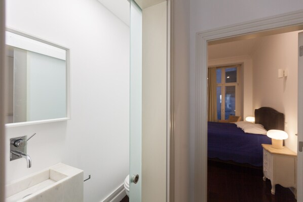Habitación de hotel con pared blanca, baño con lavamanos y espejo, cama con cubrecama azul y mesita de noche con lámpara.
