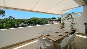 Une terrasse extérieure avec une table en bois disposée pour quatre personnes, entourée de chaises blanches, sous une tonnelle blanche.