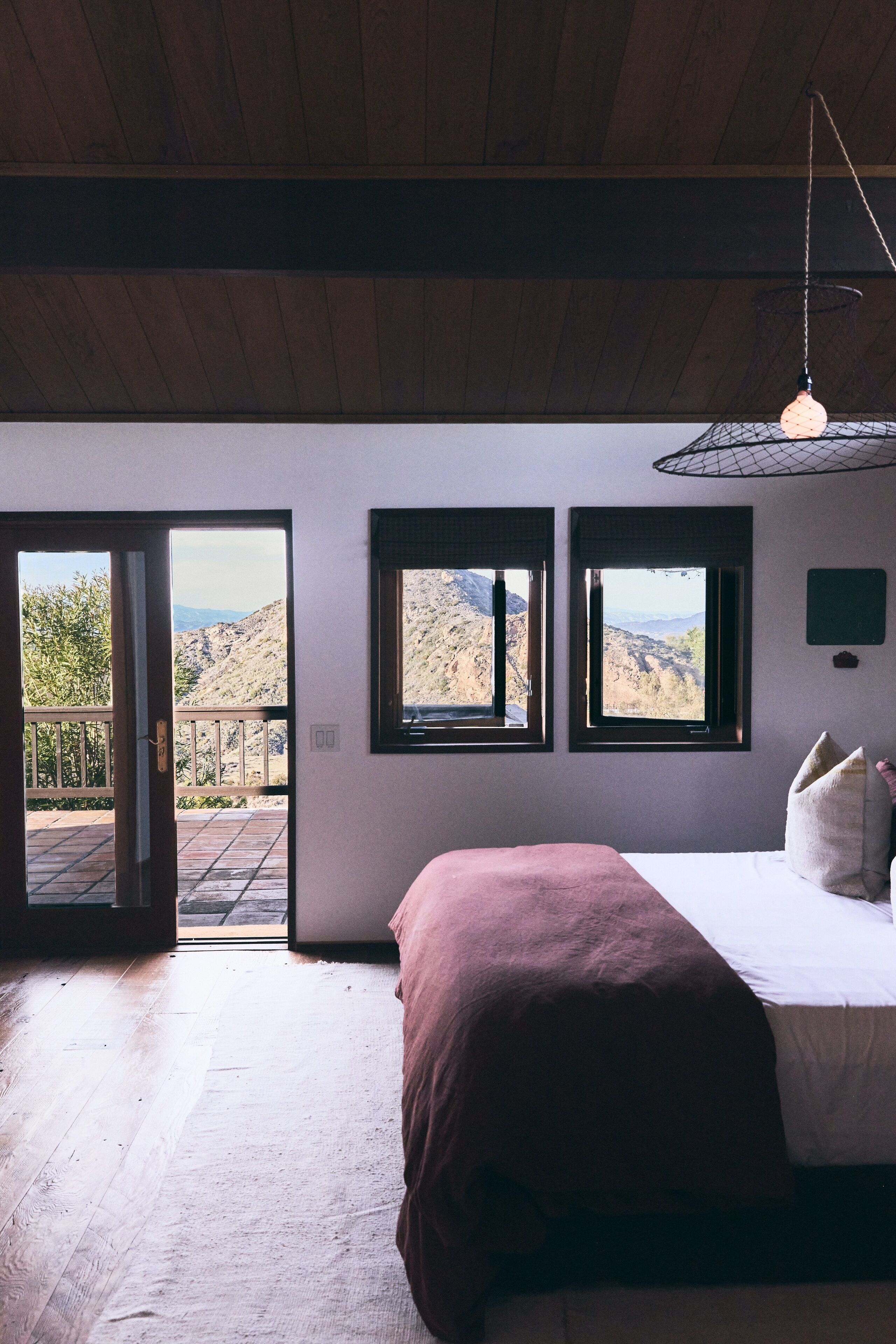 Quarto com cama, piso de madeira e três janelas com vista para o exterior.