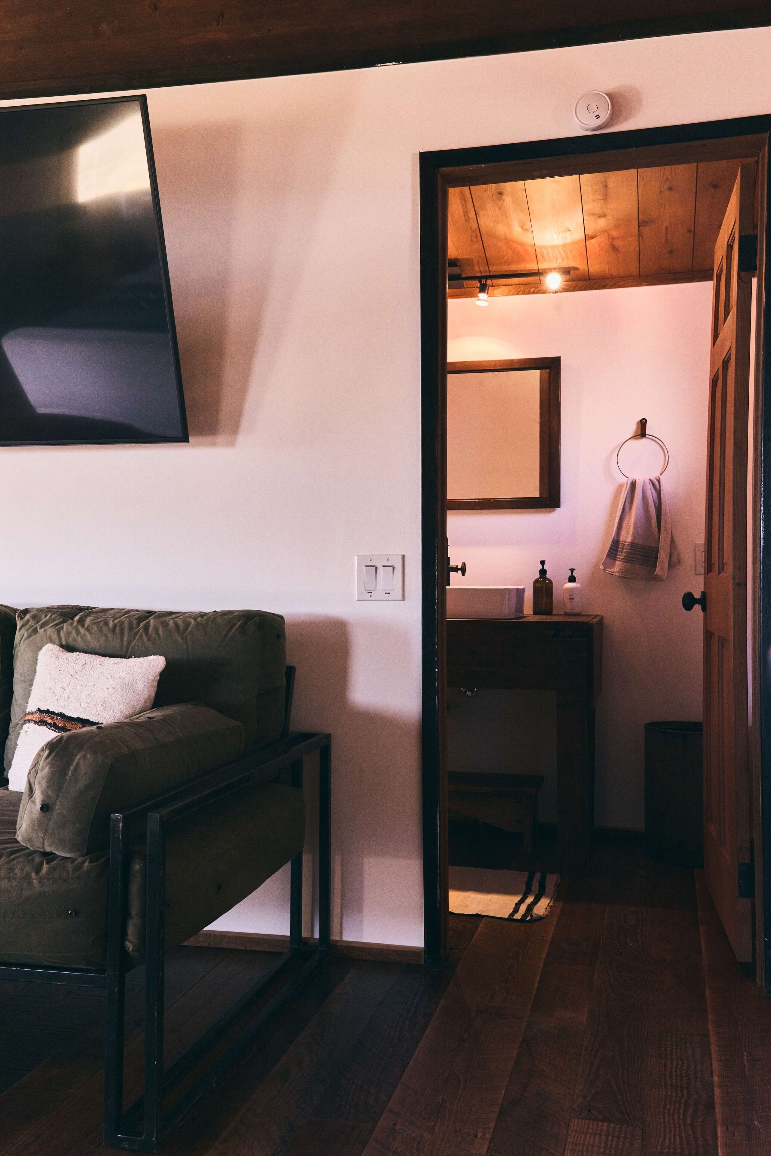 Una habitación con piso de madera, un sofá con un cojín, televisor montado, un baño con lavamanos y toallero, y techo de madera.