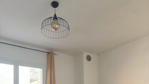 A modern living room with a sofa, a wooden table, a hanging light fixture, a potted plant, and a window with curtains.