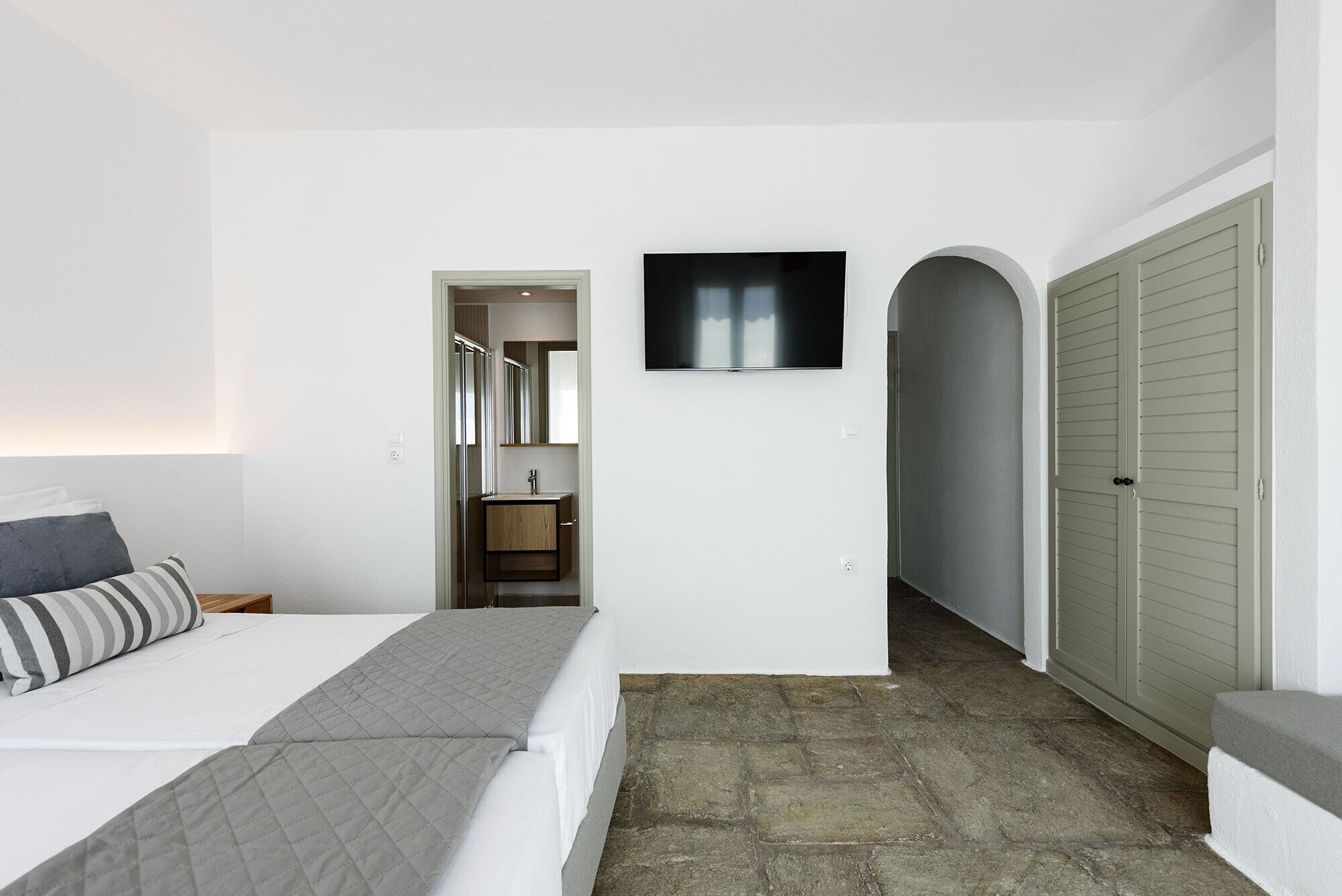 Un dormitorio con cama, televisor montado en la pared, baño con espejo y lavamanos, y armario con puertas correderas.