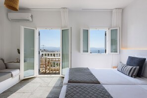 Quarto de hotel com cama, sofá, varanda com mesa e cadeiras, e vista para o mar e edifícios.