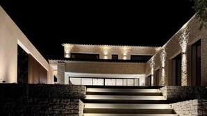 Een modern huis met een stenen gevel, een reeks verlichte treden die leiden naar een overdekt terras, en een groot raam op de bovenverdieping.