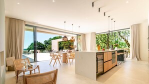 Een moderne keuken met een centraal eiland, grote ramen en een eethoek met rieten meubels.