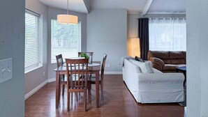 A modern living room with a dining area, featuring a wooden dining table, chairs, a white sofa, and a large window with blinds. - The PennHaus - Peaceful Home Near Lake CDA & Shops (Coeur d'Alene)