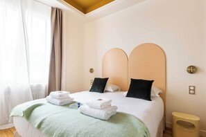 Cama bem arrumada com toalhas dobradas e um cobertor verde, uma mesinha de cabeceira e uma tomada na parede.