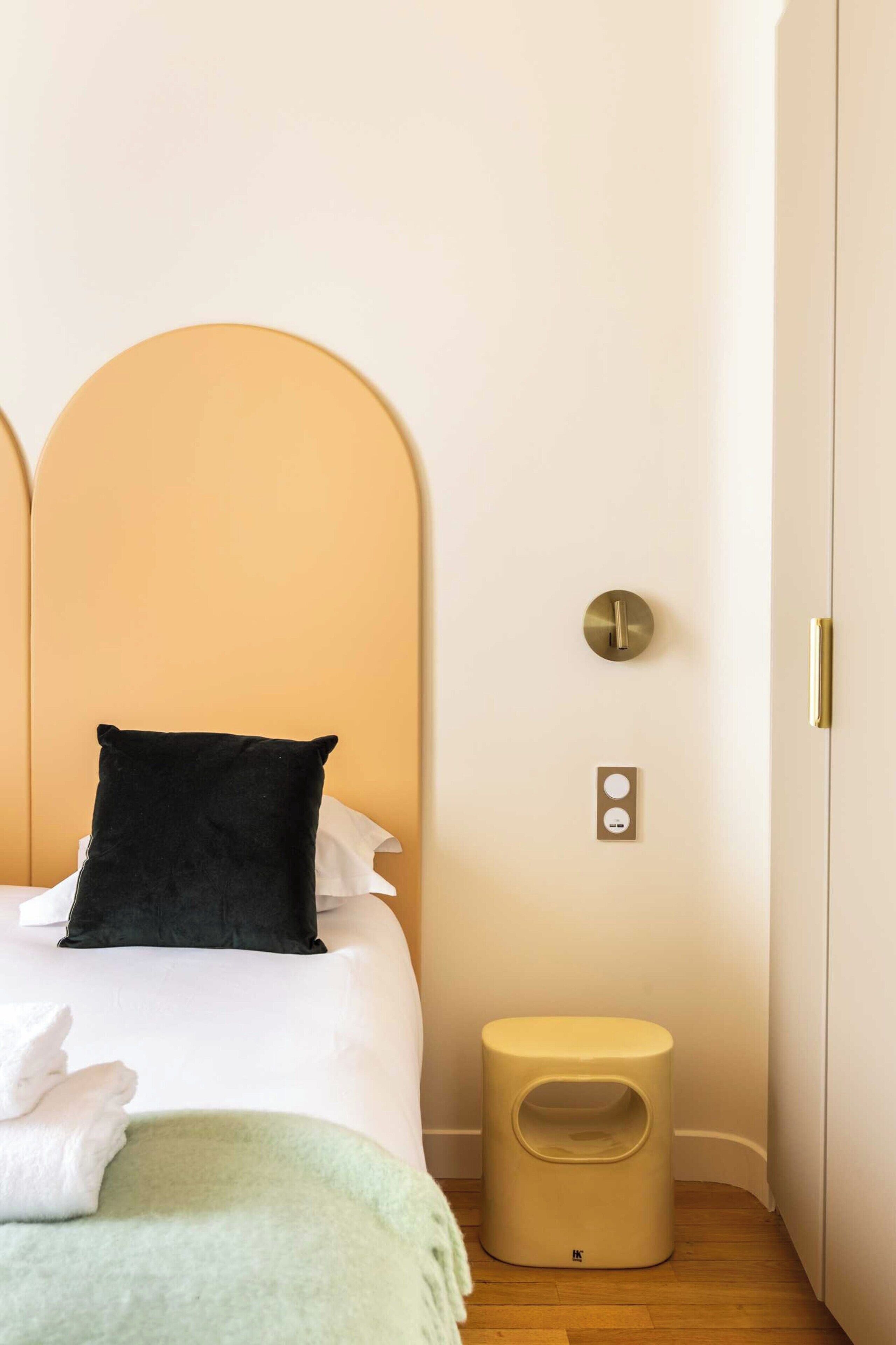 Una cama bien hecha con un almohadón negro, una mesita de noche pequeña y un interruptor de luz en la pared.