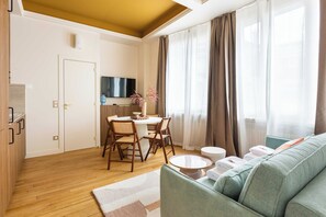 Uma sala de estar moderna com um sofá verde, mesa de jantar de madeira e cadeiras. Há uma TV fixada na parede e uma janela com cortinas.
