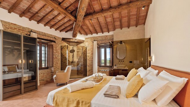 Un dormitorio con una cama grande, techo de madera, paredes de piedra, puerta de cristal y un baño con espejo.