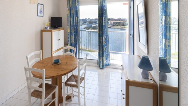 Un coin repas avec une table ronde, des chaises blanches, un téléviseur posé sur un meuble et une vue sur un plan d’eau, visible à travers de grandes fenêtres ornées de rideaux bleus.