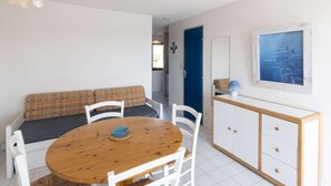 Une pièce avec une table ronde en bois, des chaises blanches, un canapé à motifs, une porte bleue, un meuble blanc et un tableau encadré au mur.