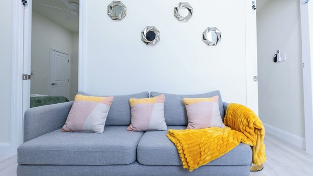 Een grijze bank met meerdere kussens, een gele plaid en decoratieve zeshoekige spiegels aan de muur.