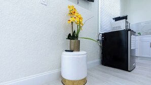 Een moderne keuken met een witte koelkast, een magnetron en een spoelbak. Op een witte kruk staat een potplant met gele bloemen.