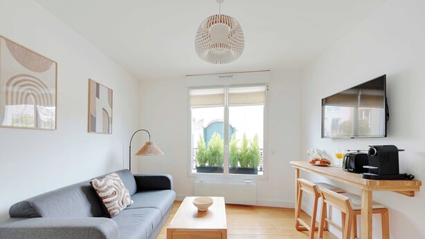 A modern living space with a grey sofa, a wooden coffee table, a kitchen area with a TV, and a large window with a view of greenery. - Charming Apartment - 1br-4p- Suresnes (Suresnes)