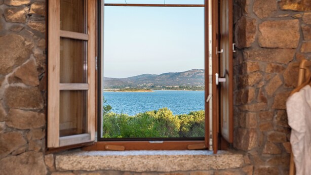 Vista attraverso una finestra aperta con pareti in pietra, su un corpo d'acqua e dolci colline in lontananza.