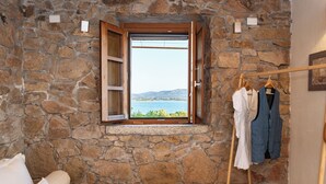A stone-walled room with a bed, a window with an open shutter, and a clothing rack with garments. - L'isola che non c'è NG Collection (Olbia)