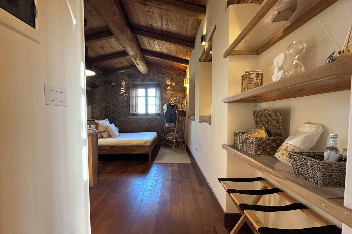 Un pasillo estrecho con suelo de madera que conduce a un dormitorio con pared de piedra y una ventana. A la derecha, hay estantes con cestas y botellas.