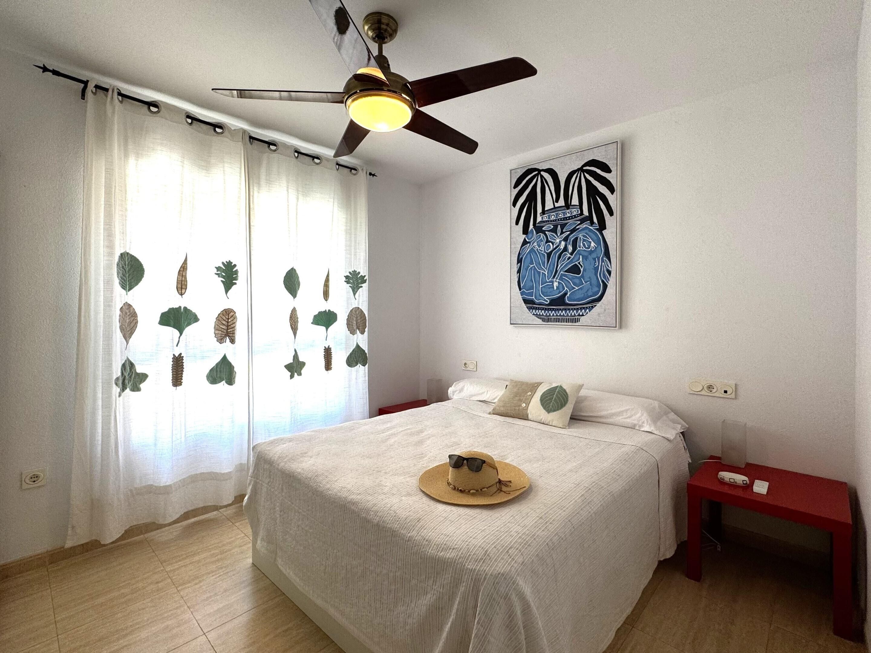 A bedroom with a bed, a ceiling fan, a wall-mounted artwork, a nightstand, and a window with curtains.