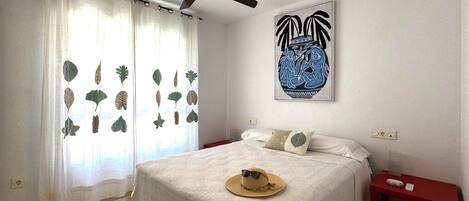 A bedroom with a bed, a ceiling fan, a wall-mounted artwork, a nightstand, and a window with curtains.
