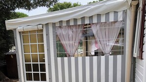 A small, single-story structure with a white exterior and striped awning. It features a front door with glass panels, a set of wooden steps leading up to the entrance, and a small porch area with potted plants and a decorative statue. - Rent MOBILHOME air-conditioned quiet terrace covered bicycles 10mn beach LA TREMBLADE (La Tremblade)