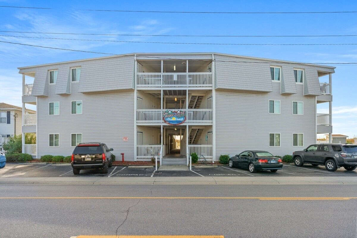 A multi-story building with a central entrance, balconies, and a parking lot with several cars.