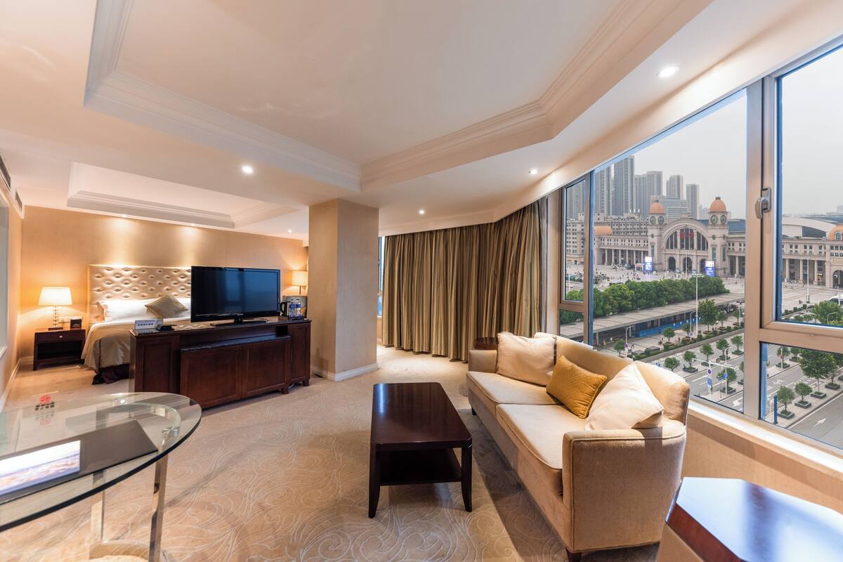 A modern hotel room with a large window, a sofa, a coffee table, a TV, and a glass table.