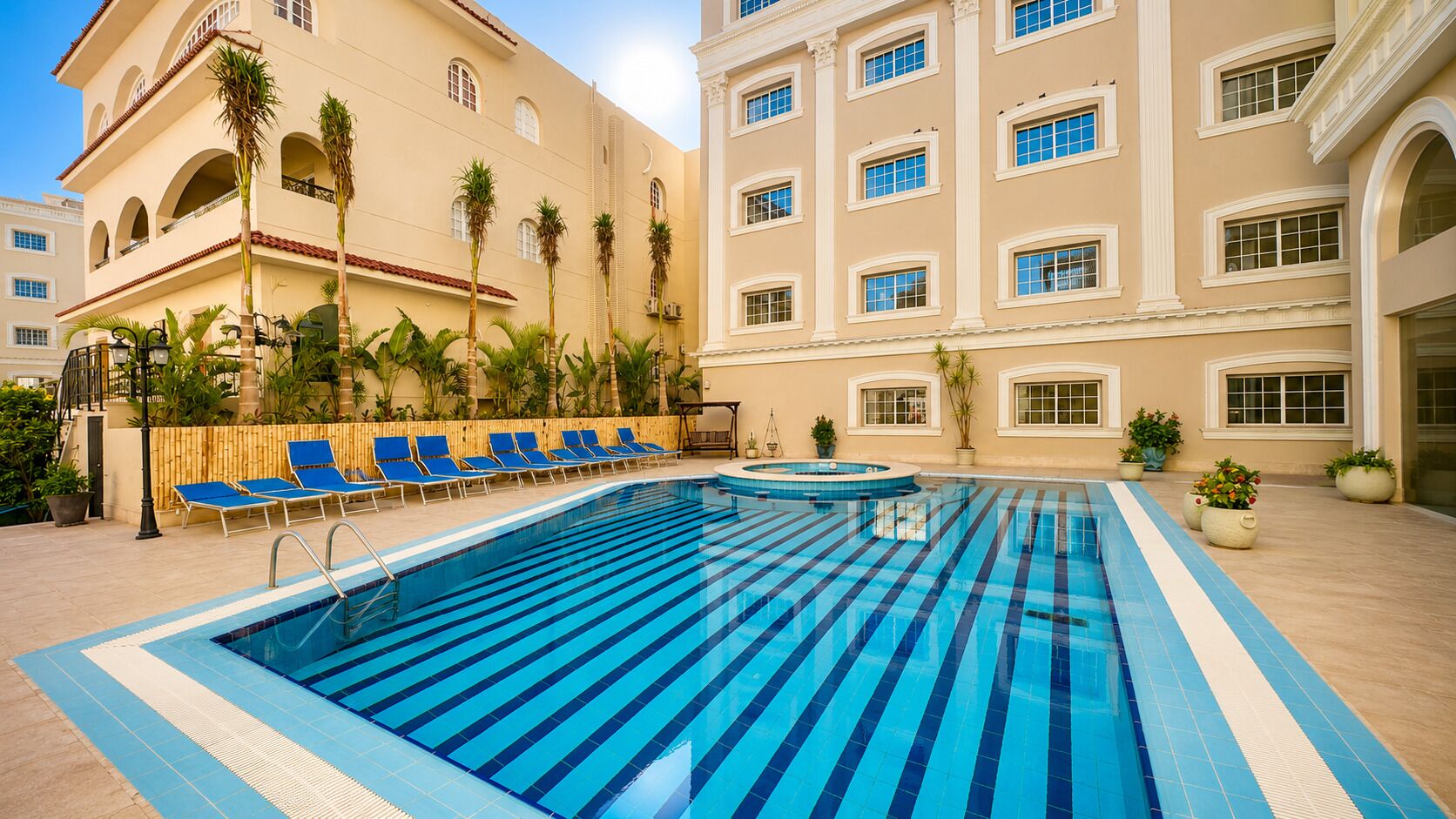 A pool area with blue-striped tiles, lounge chairs, and a building with balconies and windows.