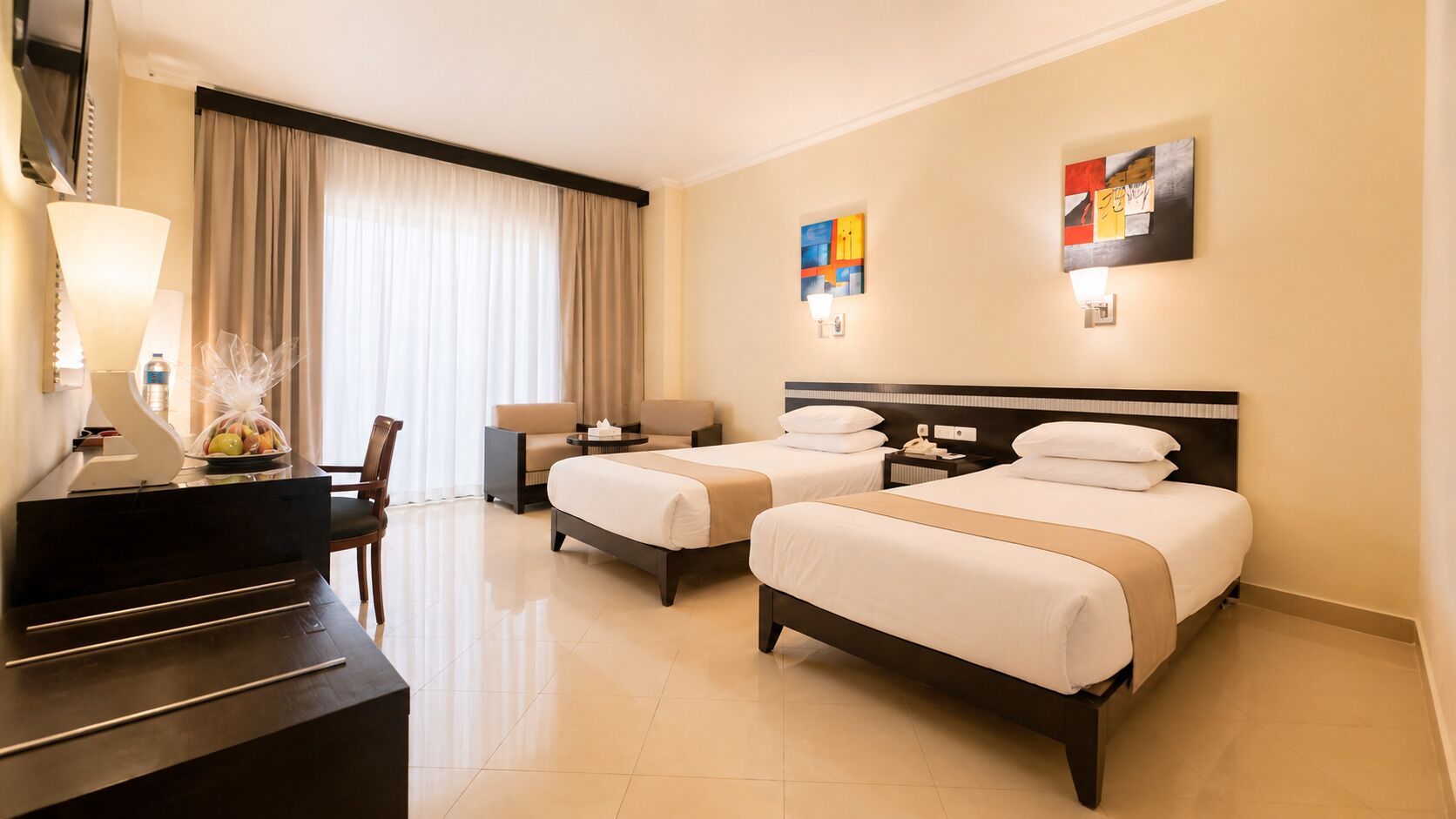 Een hotelkamer met twee bedden, een bureau, een stoel, een lamp en kunstwerken aan de muren.