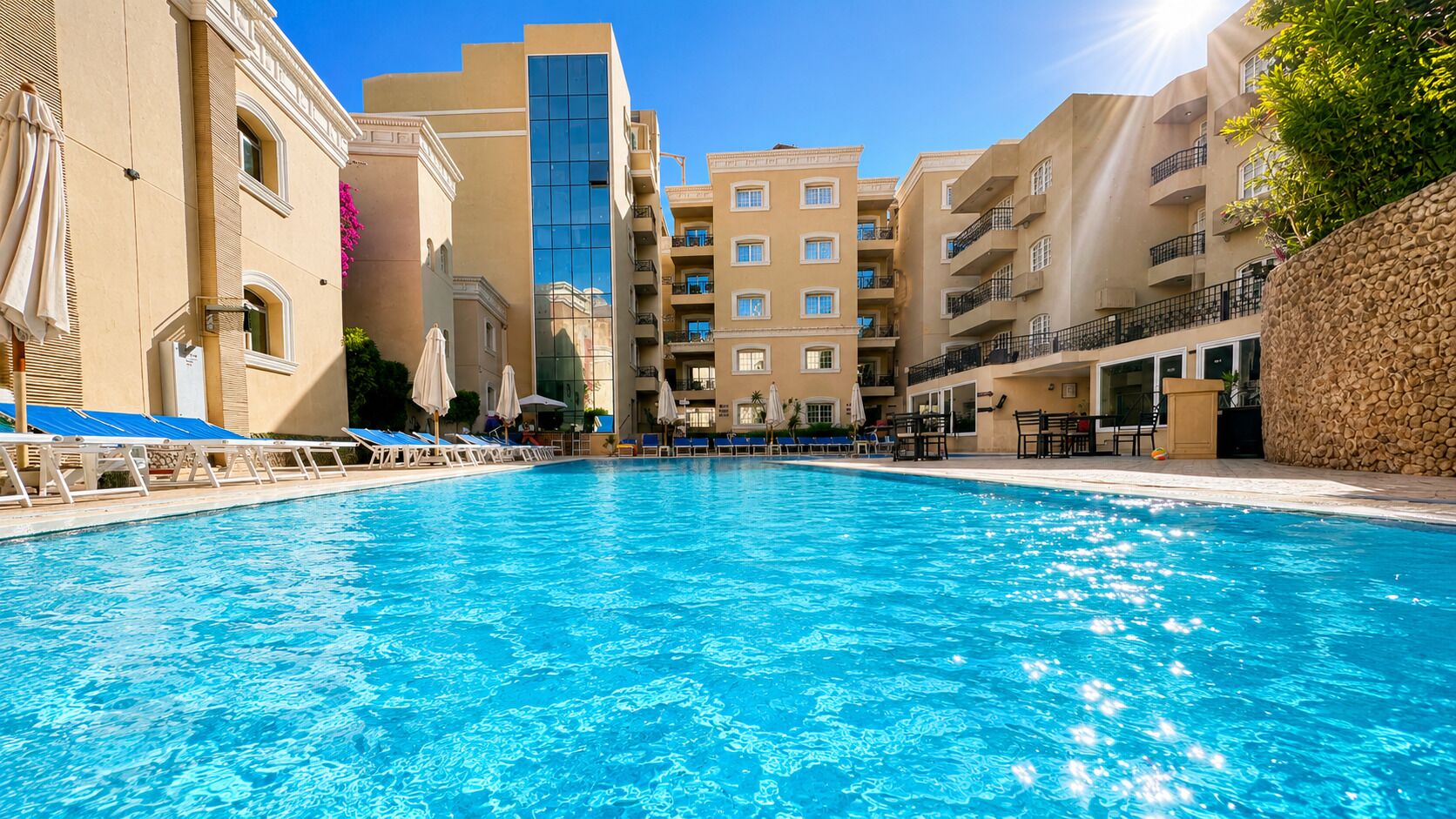 A swimming pool with lounge chairs and umbrellas, surrounded by multi-story buildings with balconies.