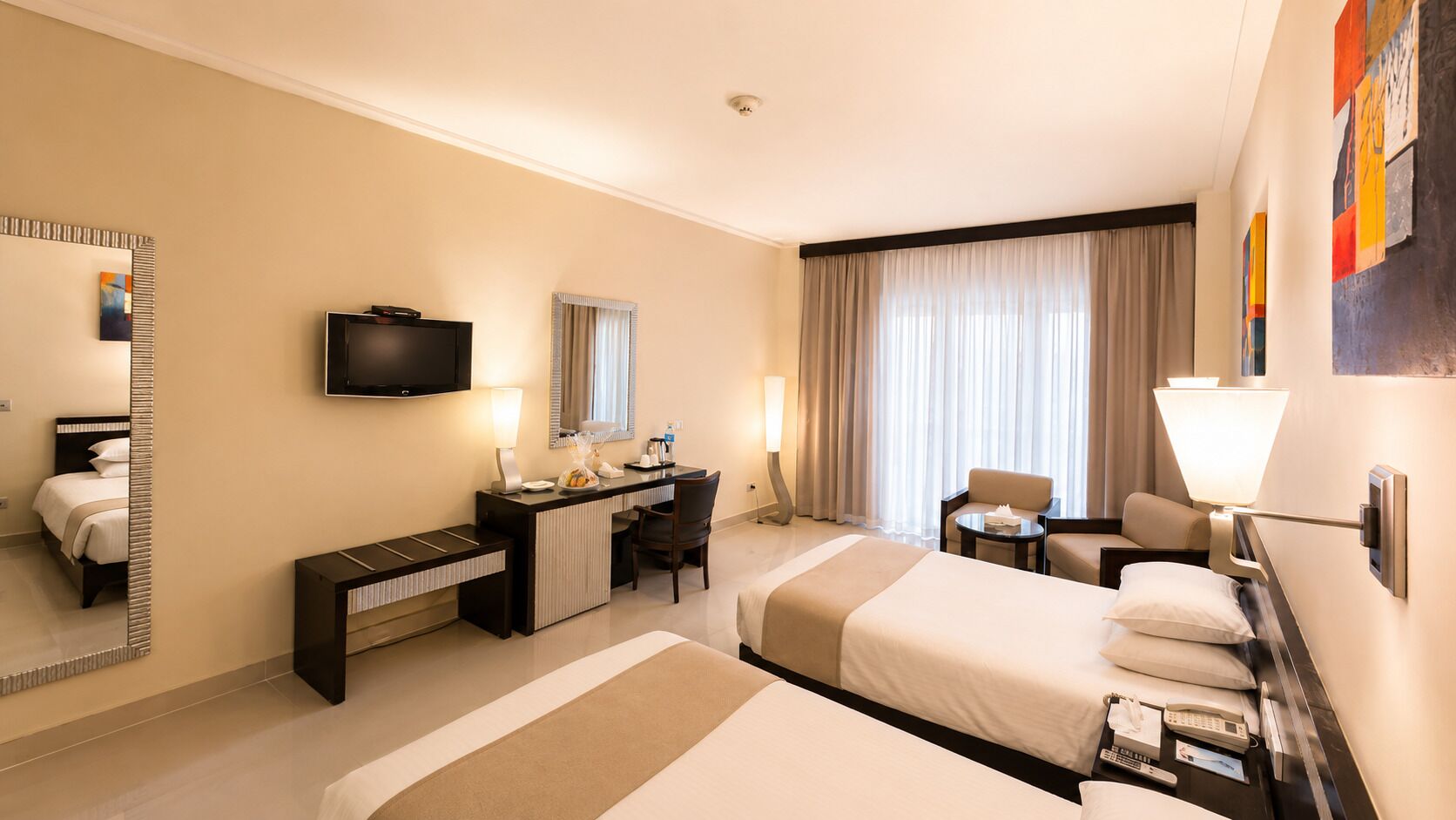 A hotel room with two beds, a desk, a chair, a TV, and a large window with curtains.