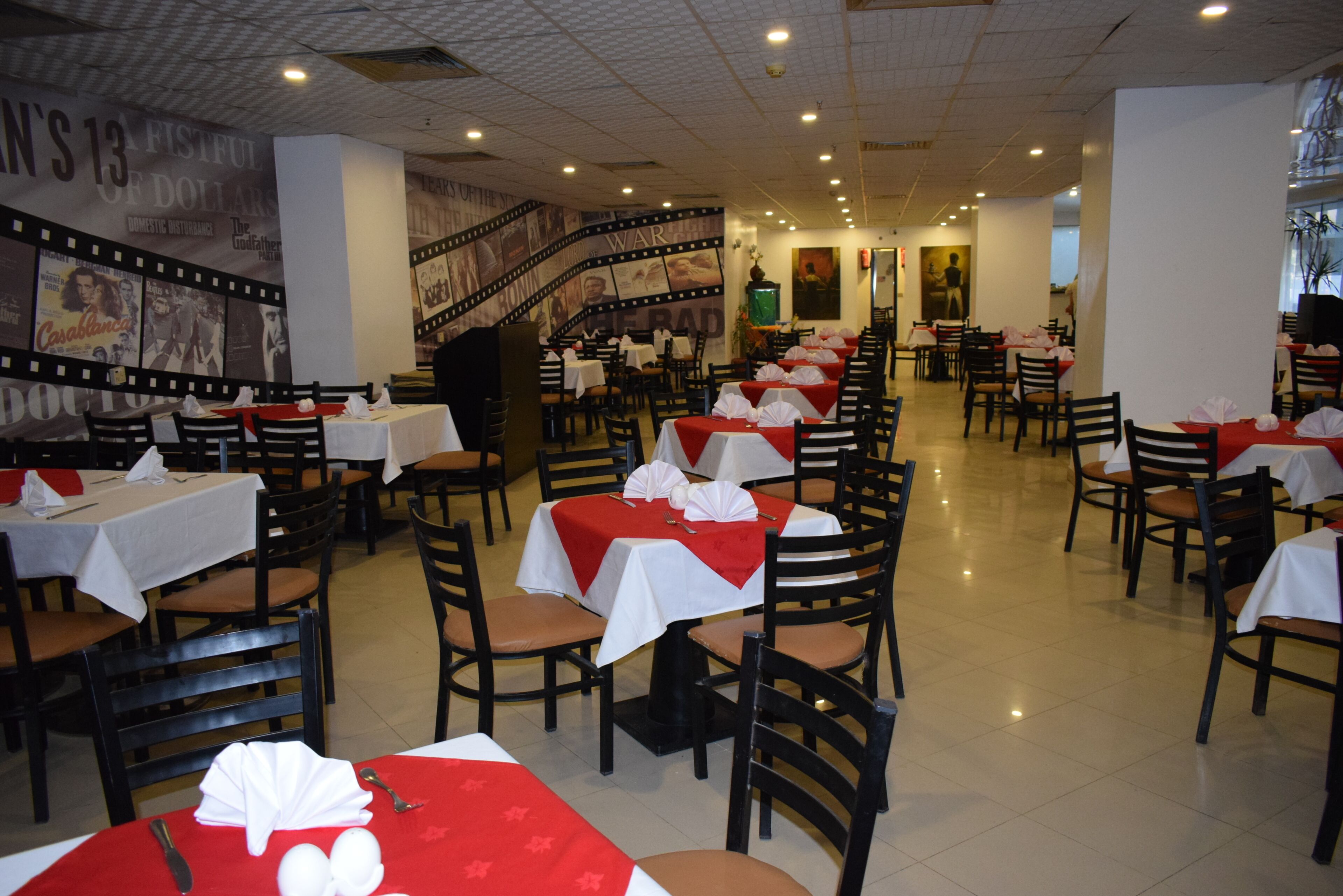 A restaurant with tables covered in white tablecloths and red napkins, wooden chairs, and a film strip mural on the wall.