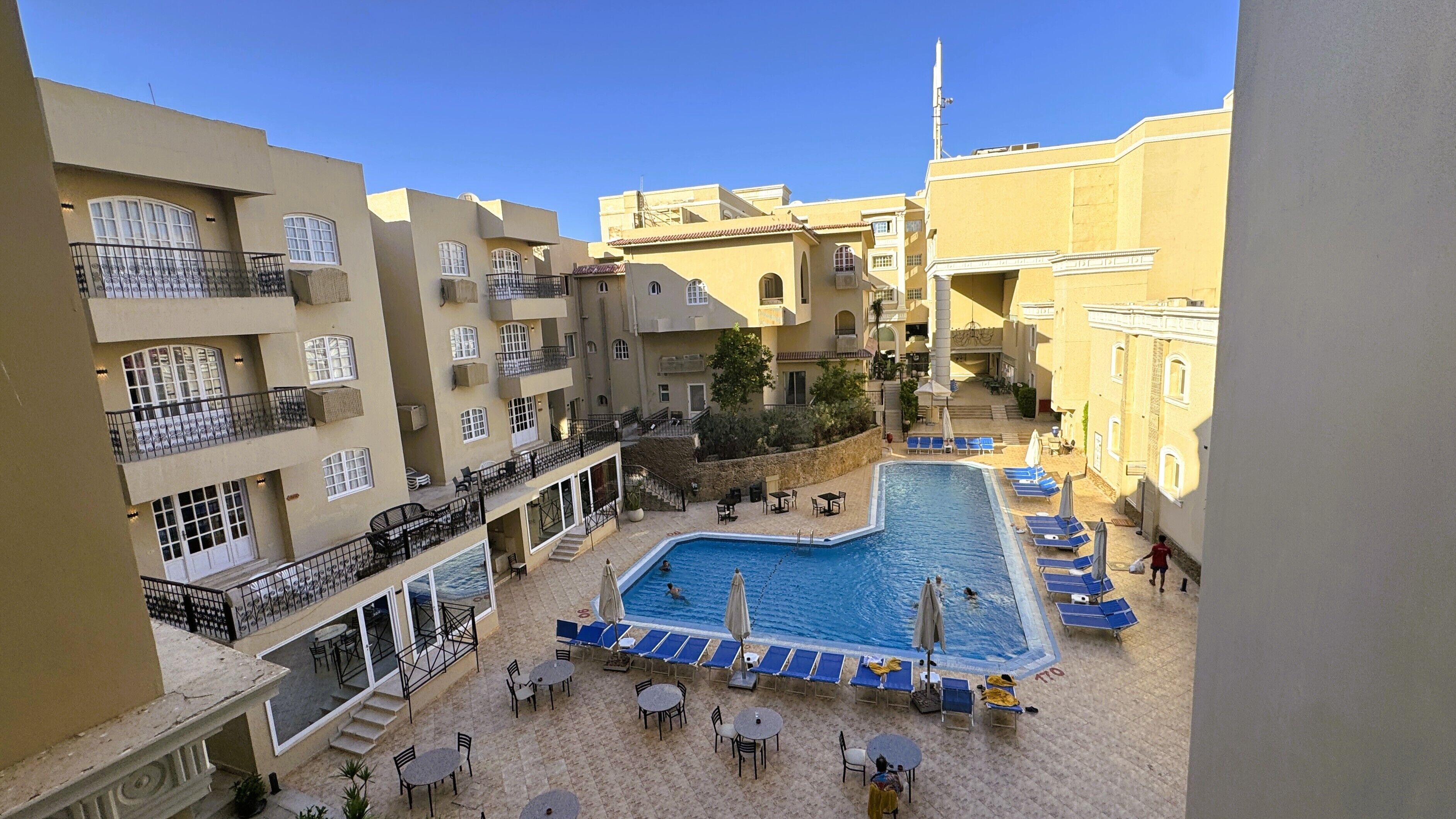 A rooftop pool area with lounge chairs and tables, surrounded by multi-story buildings with balconies.