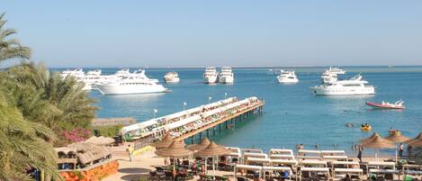 A marina with multiple yachts docked, a pier with sun loungers, and a beach area with umbrellas.