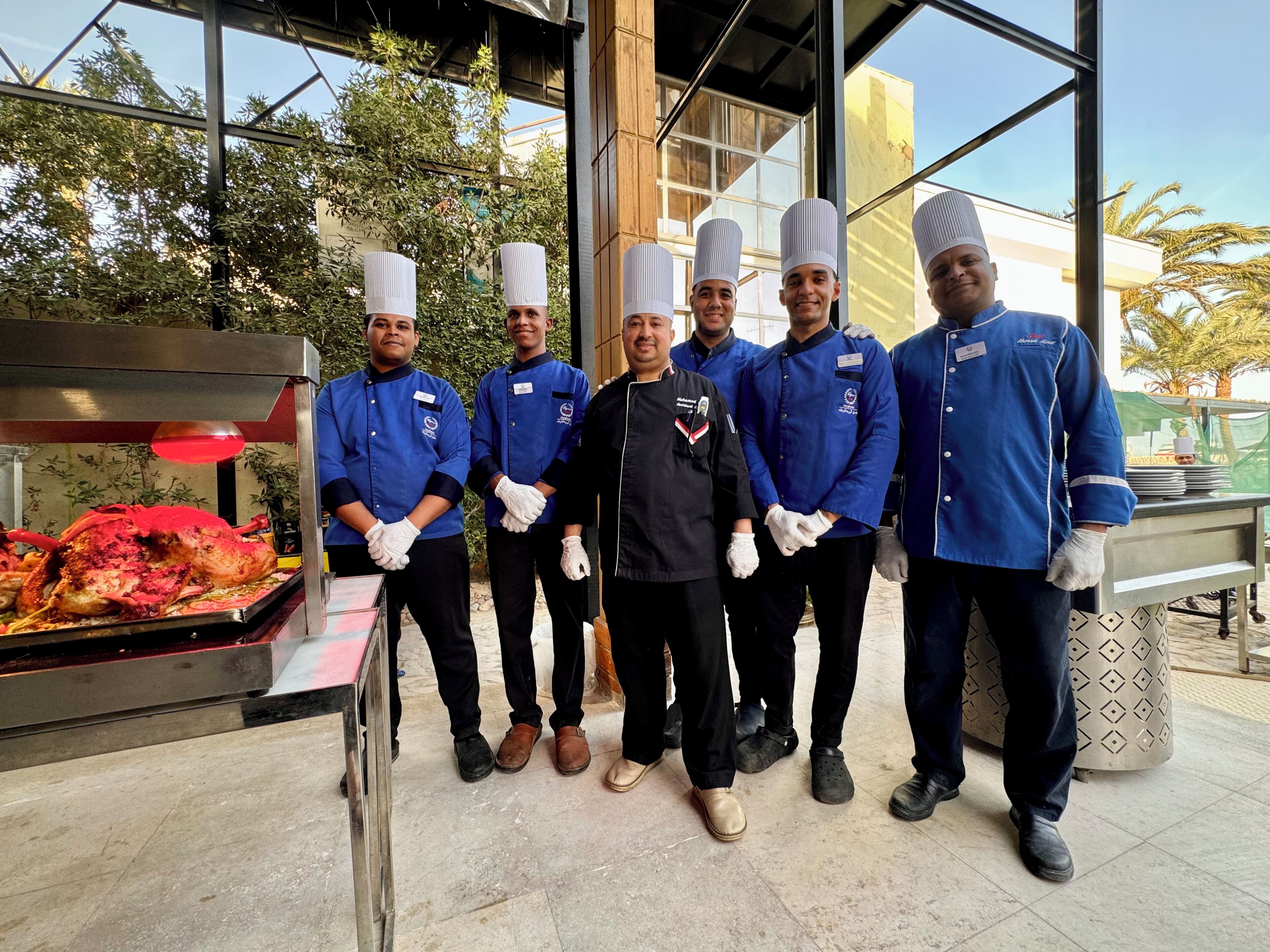 A group of people in uniform standing in front of a food display with a large roasted chicken.