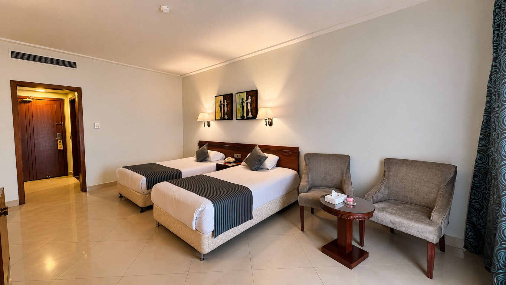Quarto de hotel com duas camas, uma cadeira, uma mesinha e obras de arte na parede.