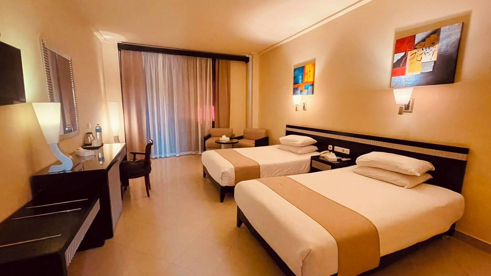 A hotel room with two beds, a desk, a chair, a small table, and a window with curtains.