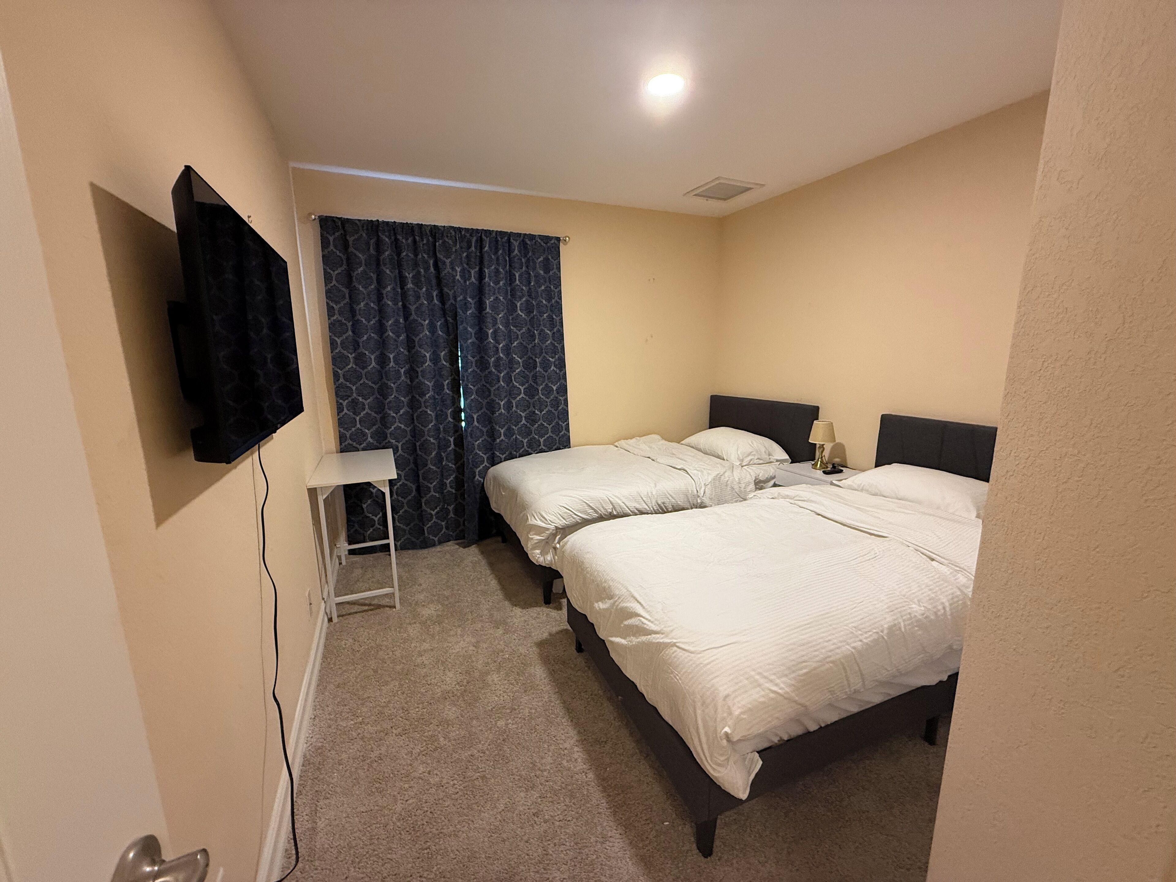 Ein Hotelzimmer mit zwei Betten, einem flachbildfernseher an der Wand, einem kleinen Nachttisch mit Lampe und einem Vorhang mit geometrischem Muster.