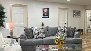 Ein Wohnzimmer mit grauem Sofa, gemusterten Kissen, einem Glastisch mit Büchern und einer Vase mit Sonnenblumen sowie zwei Türen mit Postern von Arkansas.