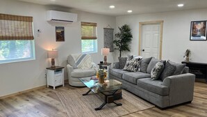 Ein modernes Wohnzimmer mit einem grauen Sofa, einem Glastisch und einem weißen Sessel. Ein kleiner Beistelltisch mit einer Lampe, ein großes Fenster mit Jalousien und eine Wandklimaanlage sind ebenfalls vorhanden.
