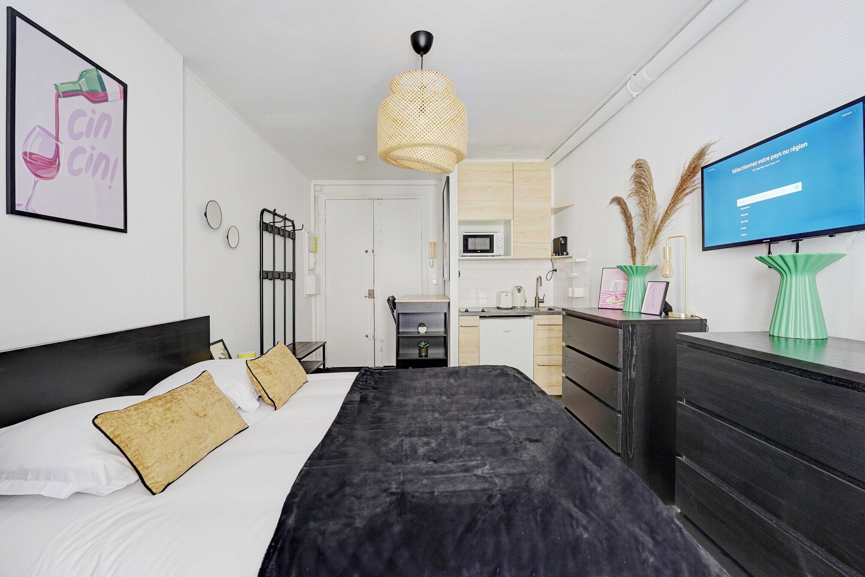 A modern hotel room with a bed, a desk, a television, and a kitchenette.