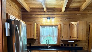 Una cocina de madera con un refrigerador de acero inoxidable, armarios de madera, una encimera negra y una ventana con cortina.