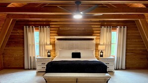 Un dormitorio con techo de madera, una cama con ropa de cama blanca, dos mesitas de noche con lámparas y una ventana con cortinas.