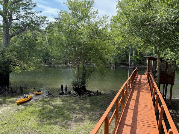 A wooden bridge leading to a dock with kayaks and a small wooden structure.