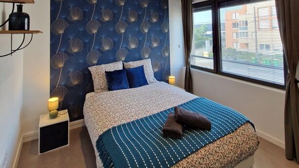 A bedroom with a large bed, a patterned headboard, a bedside table with a lamp, and a view of buildings through the window. - Chic & Central - Your Retreat in Le Mans (Le Mans)