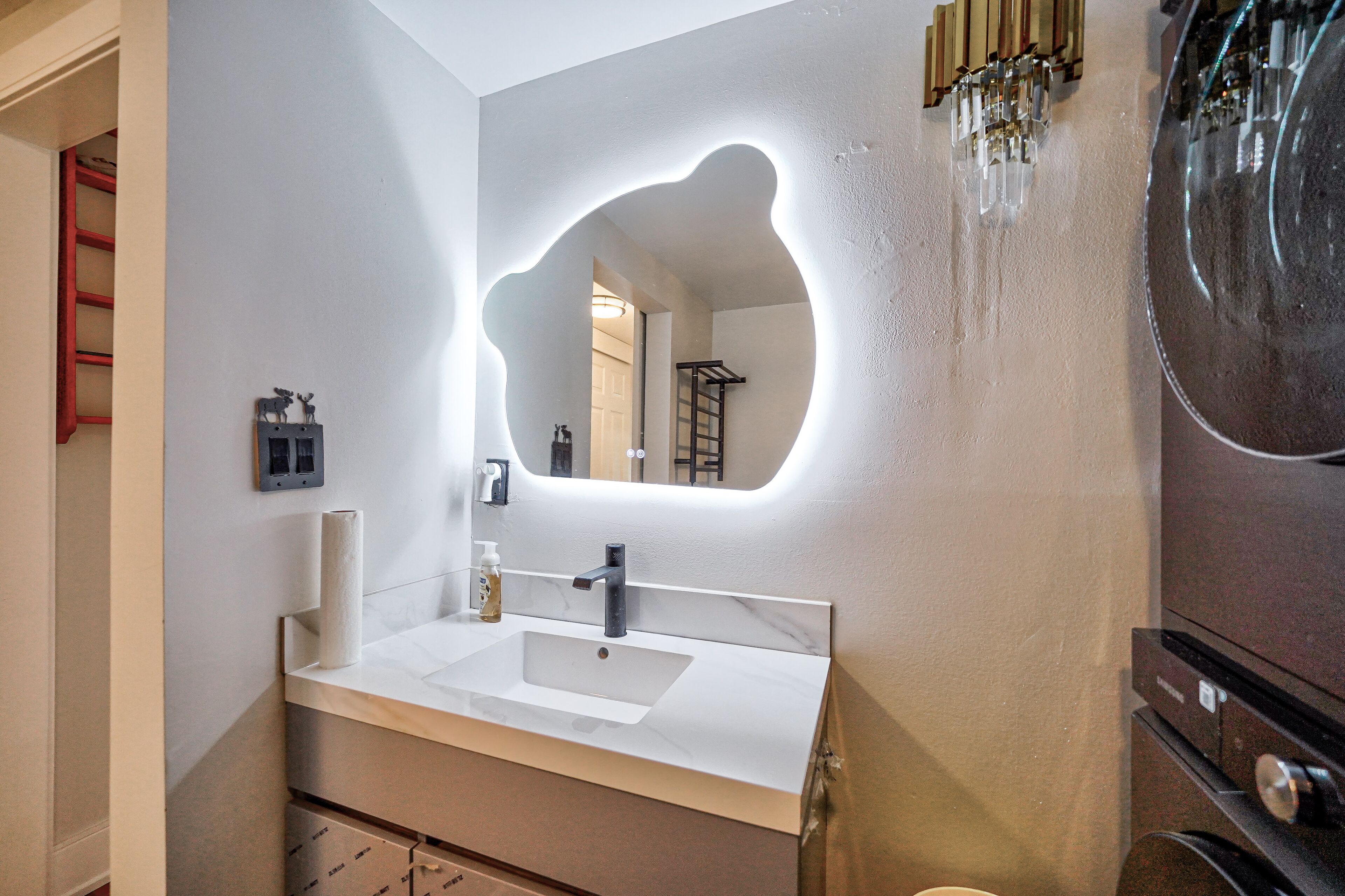 A bathroom with a white sink, a mirror with a unique shape, and a towel dispenser.