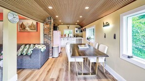 Un'accogliente zona pranzo con soffitto in legno, una cucina sullo sfondo e un'ampia finestra vista sul verde.